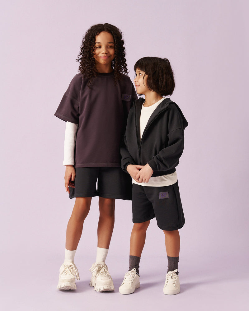 A girl and a boy side by side in ON7 tees, zip hoodie and shorts in dark tones